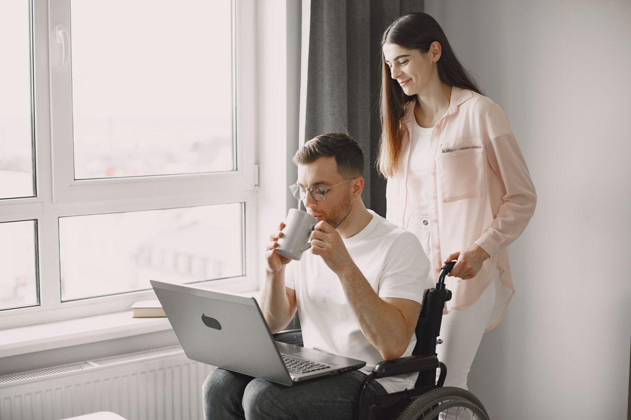 Mastering the First Impression: Your intriguing post title goes here A caring woman assists a man in a wheelchair as they enjoy coffee and work on a laptop indoors.